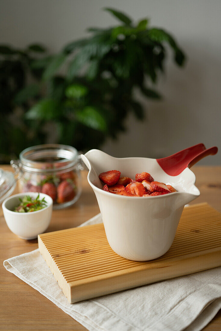 Köstliche, frisch geschnittene Erdbeeren in einer Glas- und Keramikschüssel auf einem Holztisch mit Stoff während der Zubereitung von Marmelade in einem Raum