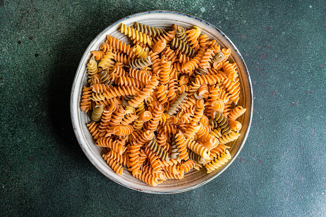 Top view of raw uncooked fusilli pasta made from wheat, beetroot and spinach in the bowl on dark green background