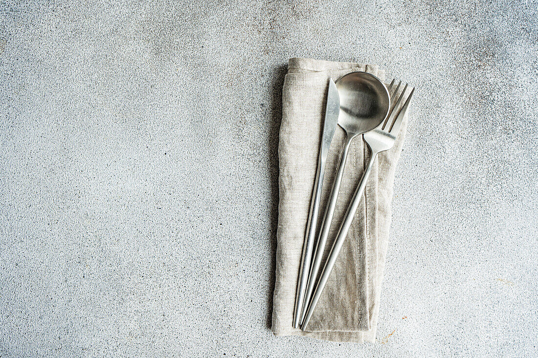 Besteckset auf grauer Oberfläche vor rauem, strukturiertem Hintergrund mit Handtuchserviette und trockenem Löffel in heller Küche