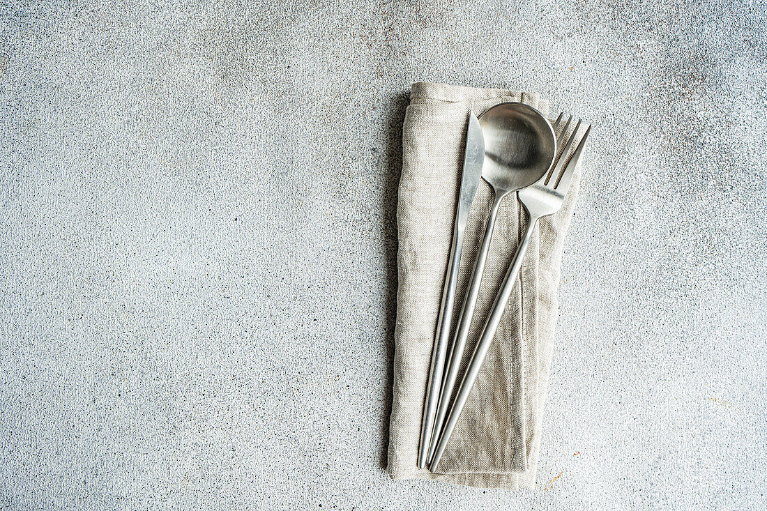 Set of cutlery placed on gray surface near rough textured background with towel napkin and dry spoon in light kitchen