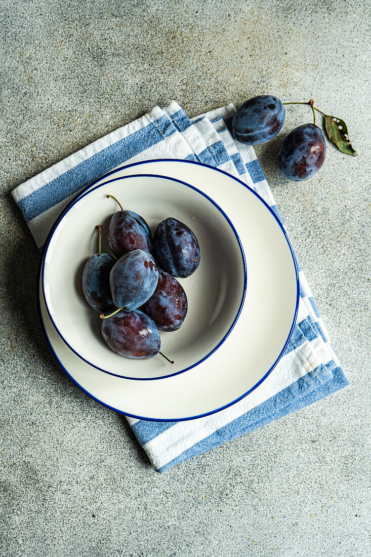 Top view of ripe plums served in ceramic bowl on striped napkin against gray surface
