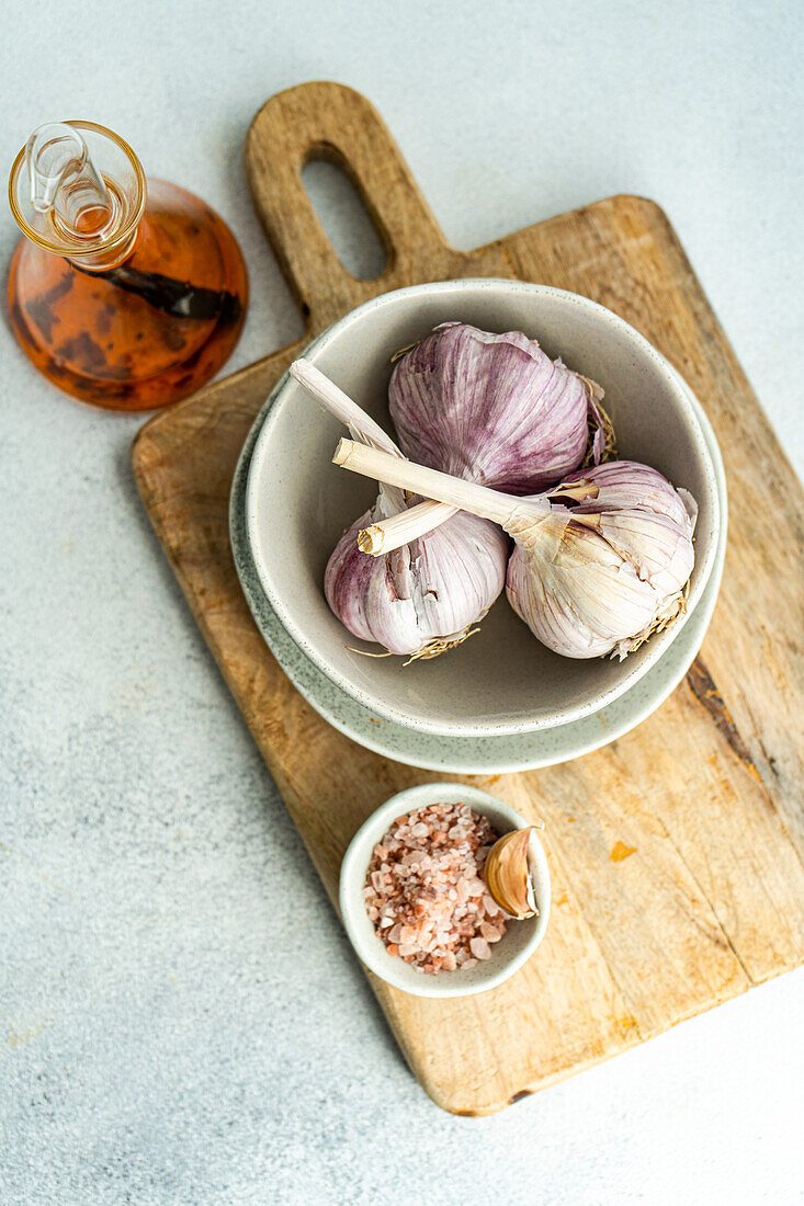 Top view of raw garlic bulbs nestled in a ceramic bowl on a rustic wooden board with an amber glass bottle nearby and a small bowl of coarse salt set against a muted gray backdrop