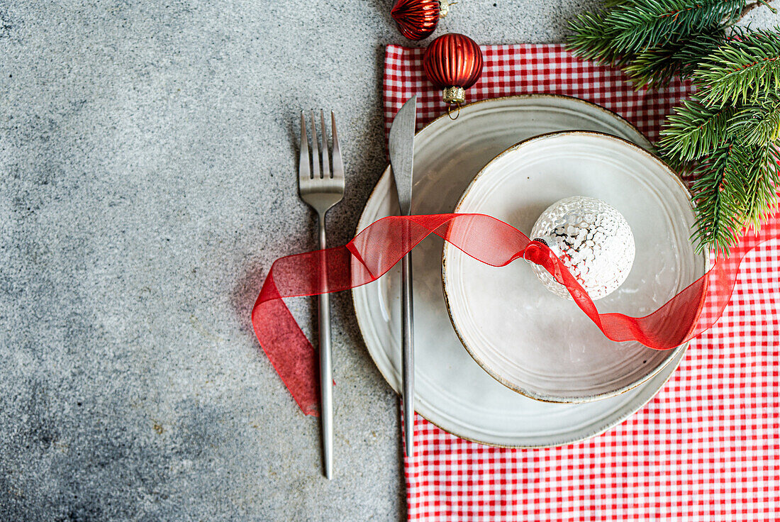From above of plates with Christmas ball decorated with ribbon and fork and knife placed on gray table