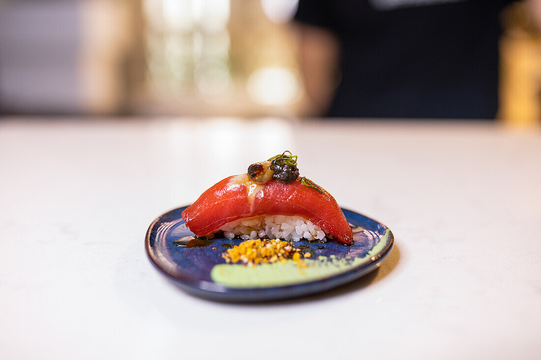 Nigiri sushi prepared with fresh tuna fish slice on top of cooked rice while garnished with sauce and served on plate on wooden table against blurred interior