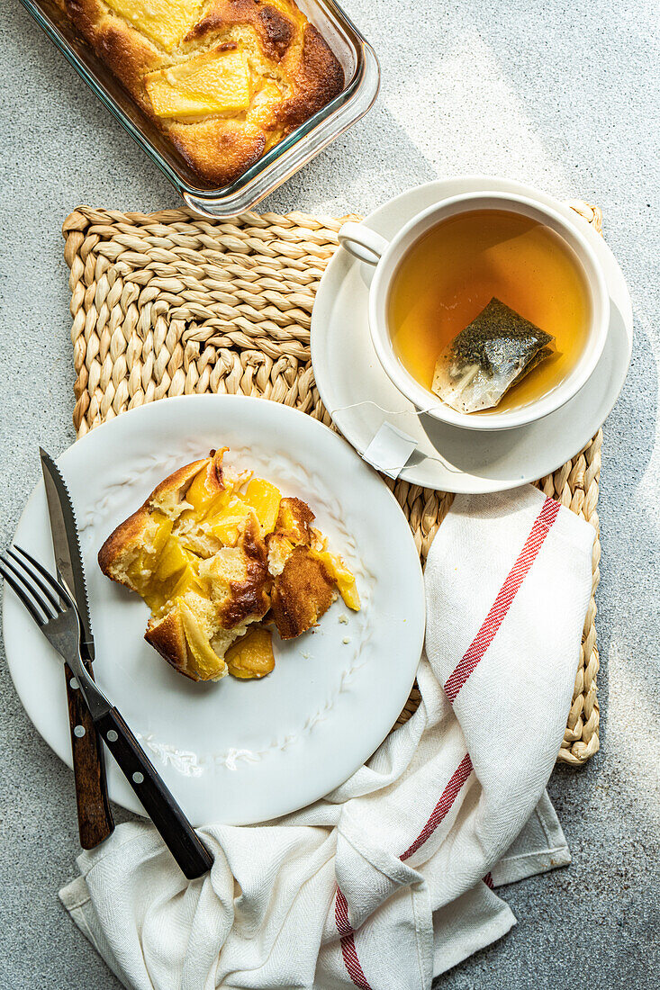 Blick von oben auf ein saisonales Stück Pfirsichkuchen, serviert auf einem Teller mit Gabel und Messer und einer Tasse Tee auf dem Tisch neben einer Serviette