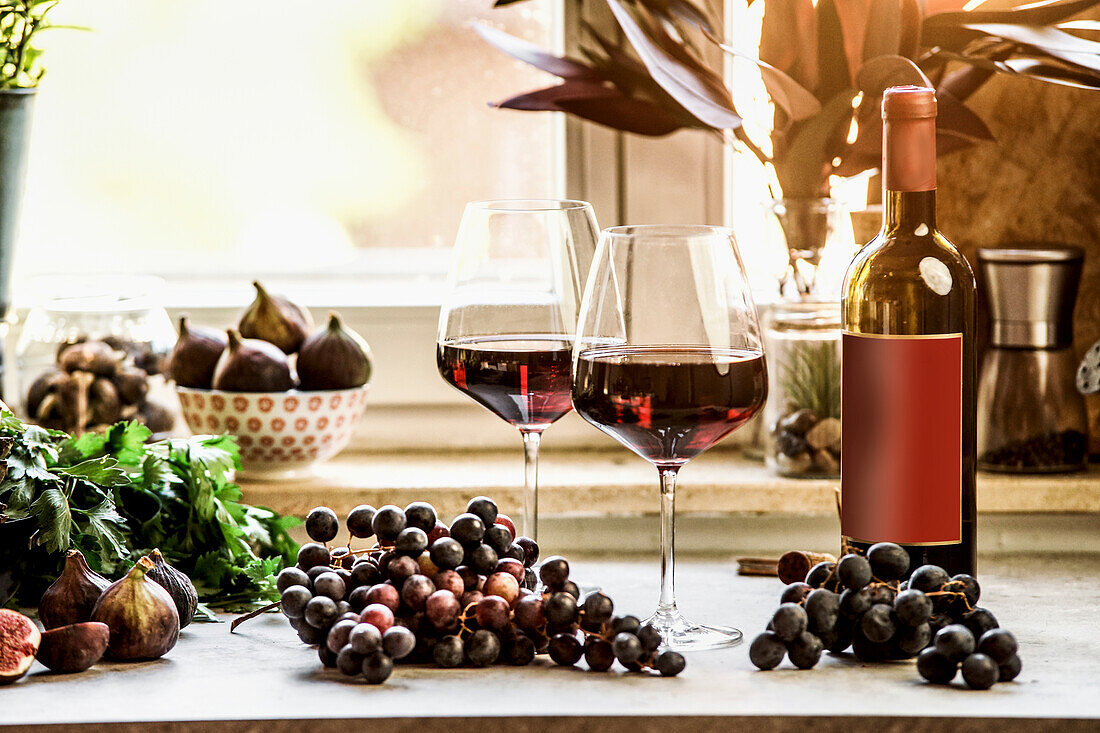 Rotweingläser mit Trauben und Weinflasche am Küchentisch mit Fensterhintergrund. Vorderansicht. Raum kopieren