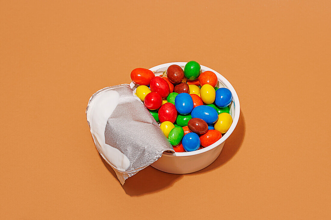High angle of opened yoghurt container filled with colored candies against orange background