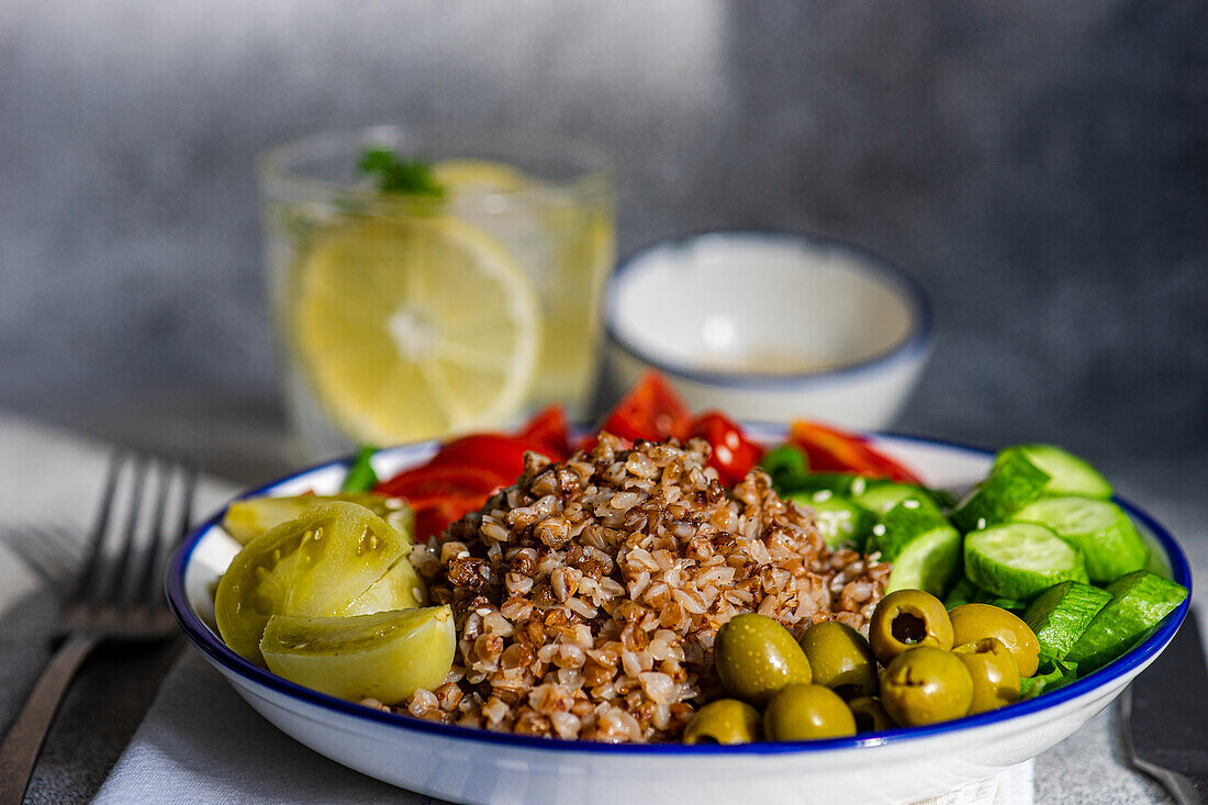 Nahaufnahme einer gesunden Mittagsschüssel mit gekochtem Bio-Buchweizen, frischer Gurke und Tomate sowie fermentierter Tomate und Oliven, serviert mit einem Glas reinem Wasser mit Zitrone, Eis und Minze vor einer verschwommenen grauen Fläche