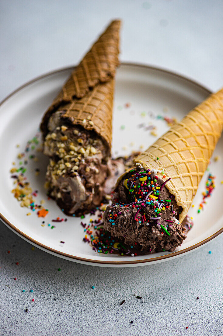 Waffle cones with coffee and chocolate ice cream with multicolored sugar sprinkles and nuts on top in a ceramic dish on a concrete background