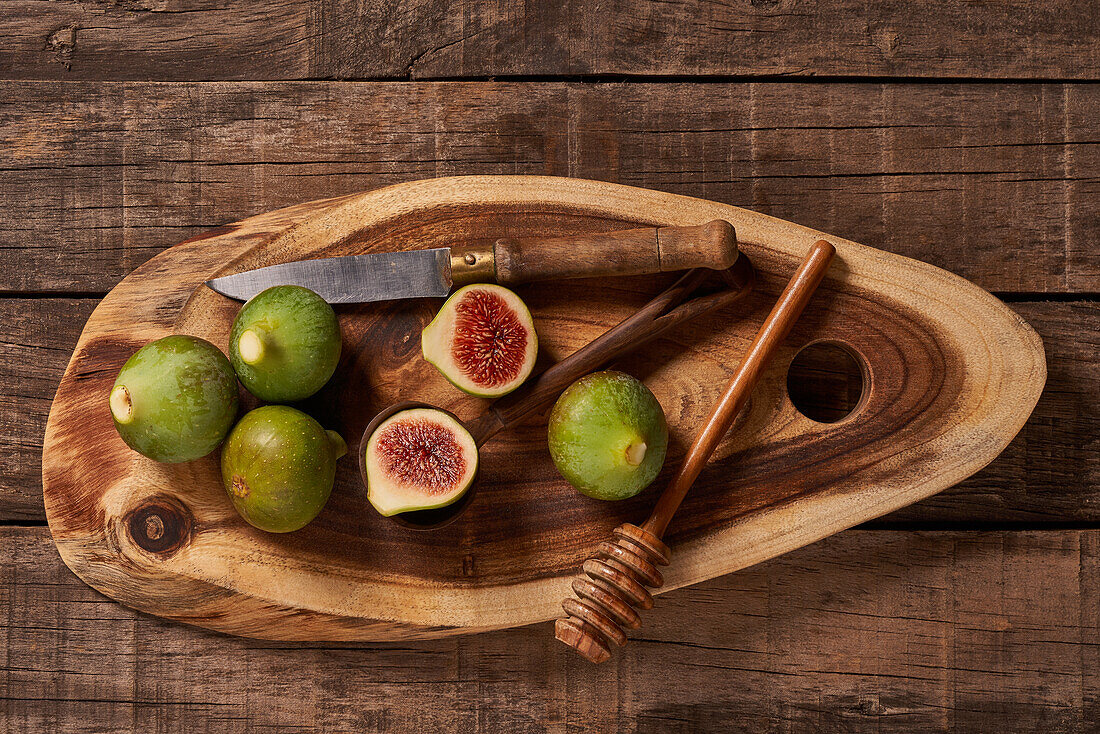 Overhead shot of a rustic wooden cutting board with fresh green figs, a half-cut fig, a honey dipper, and an old knife on a wooden background.