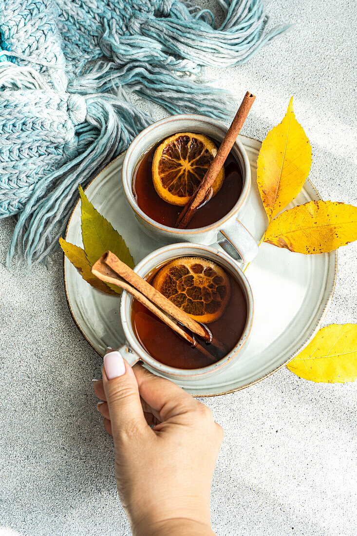 Top view of Anonymous hand reaching for one of two cups of spiced tea, accompanied by cinnamon sticks, anise, and dried orange slices, placed near radiant yellow leaves on a muted gray surface