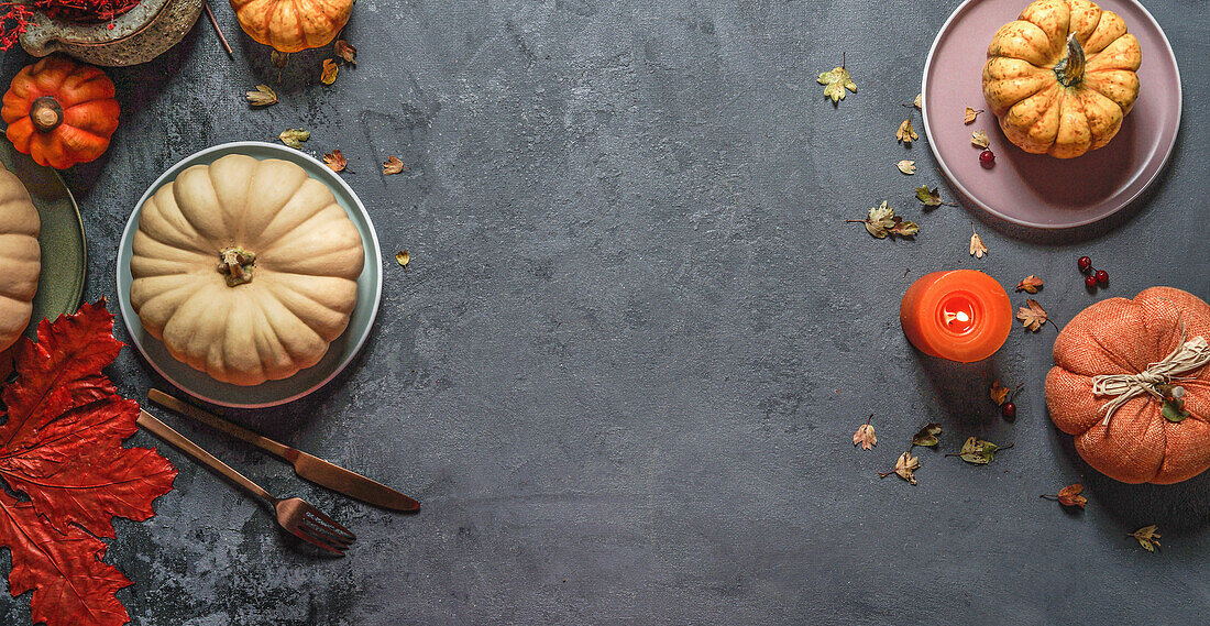 Top view of autumn background frame with various pumpkins, red fall leaves, plates, cutlery and candle at dark grey backdrop