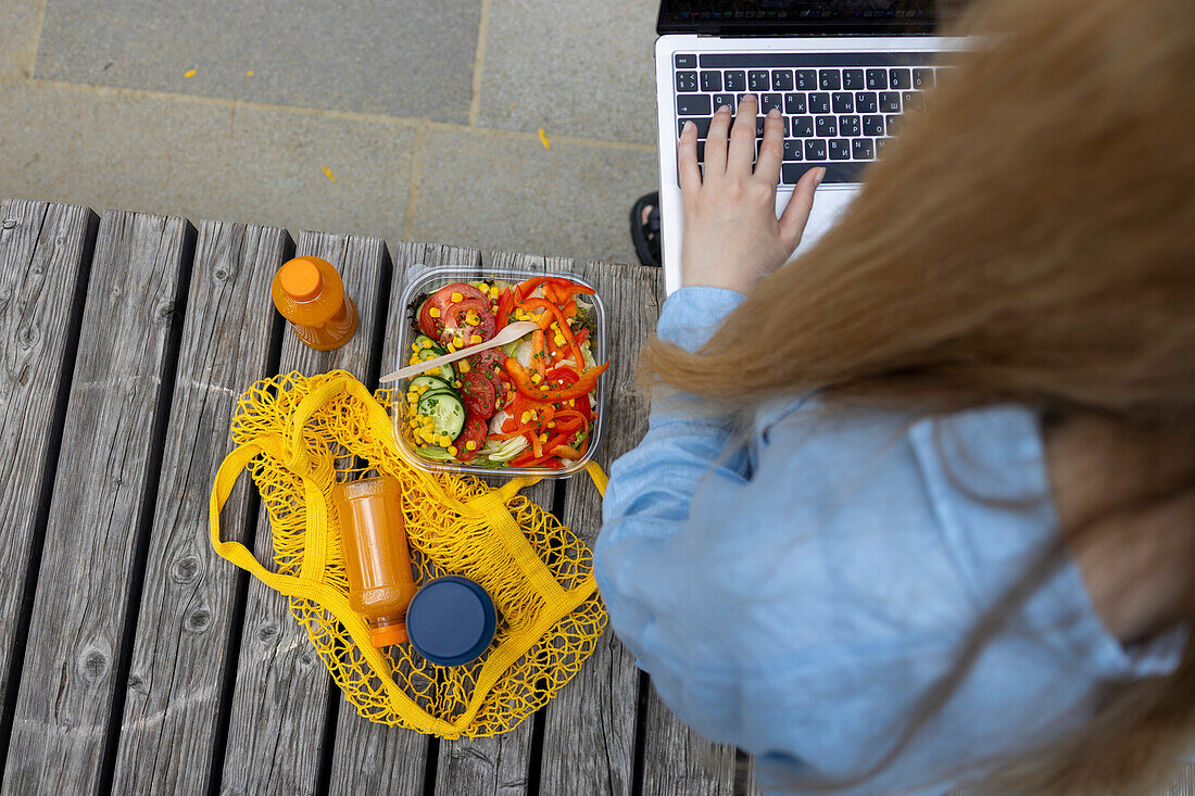 Top view of unrecognizable young blonde-haired female student writing in laptop, sitting on a bench at lunchtime outdoors.