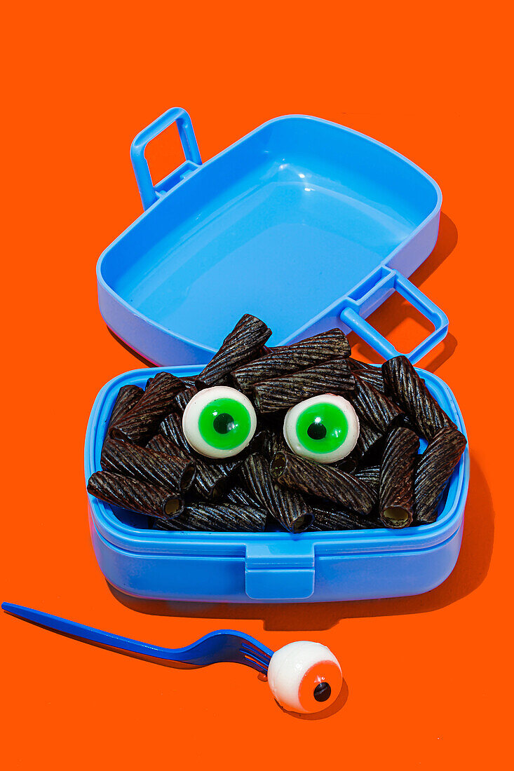 Horror-Lunch mit schwarzen Nudeln und Augen in Lunchbox auf orangem Hintergrund neben Gabel mit Auge