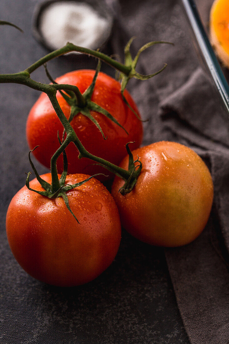 High angle closeup of fresh ripe tomatoes in stalk placed on dark plain surface with fabric indoors
