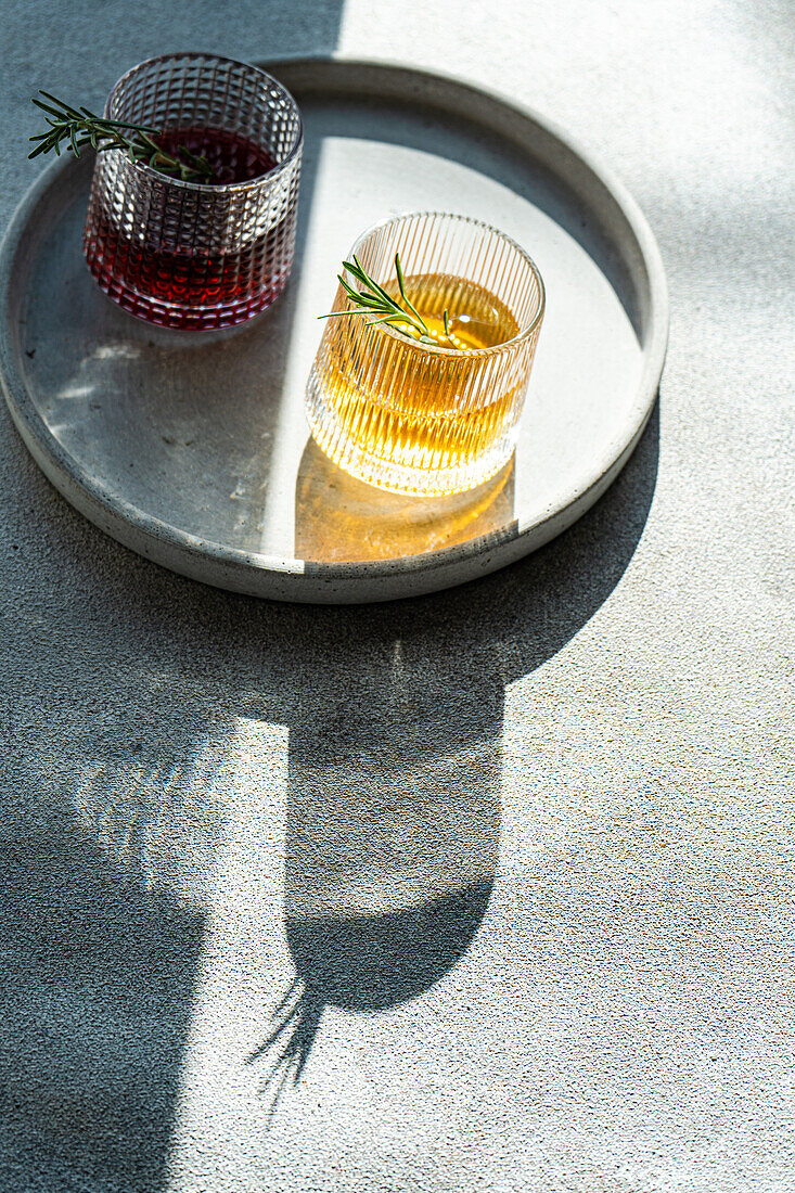 From above of artisanal cocktails with vodka cherry, and apple juice in textured glasses adorned with rosemary on a concrete surface with shadows