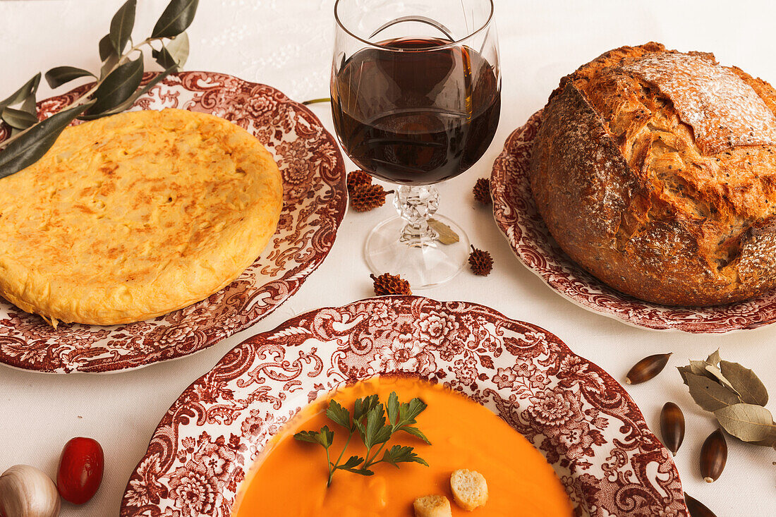 An elegant dinner setup featuring a spanish omelette and salmorejo with croutons, fresh crusty bread, a glass of red wine.