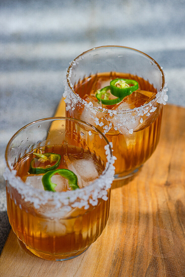 High angle of glasses with Japanese sochu liquor with mango juice and slices of Jalapeno pepper with salt on the glass edge placed on wooden table against blurred gray background