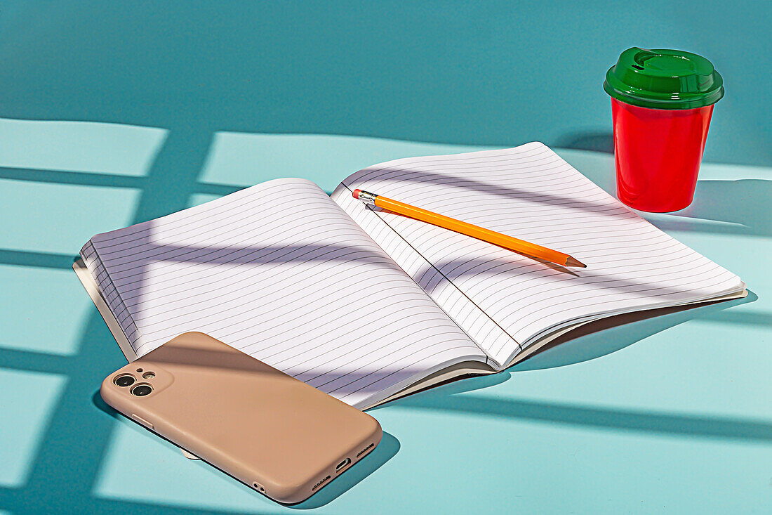 Minimalistischer roter Plastikbecher mit Kaffee und grünem Deckel zusammen mit einem Notizbuch und einem Mobiltelefon vor blauem Hintergrund