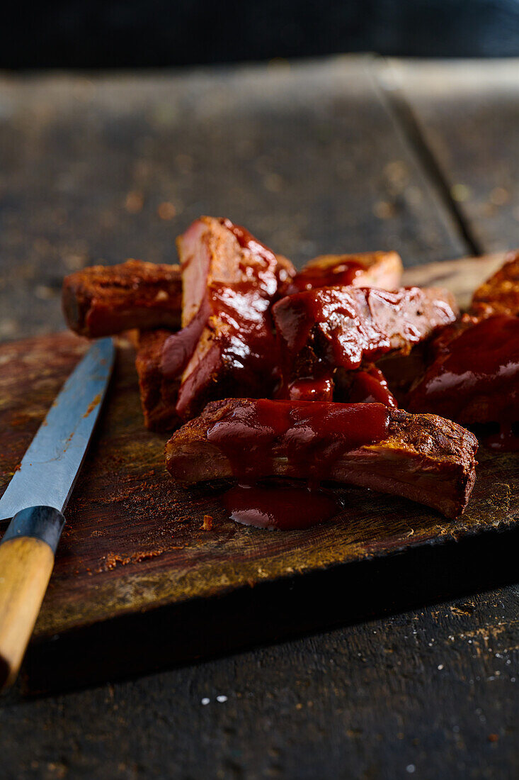 Hoher Blickwinkel auf appetitlich gegrillte Schweinerippchen, die auf einem Holzbrett in der Küche mit einem Messer vor einem unscharfen Hintergrund serviert werden