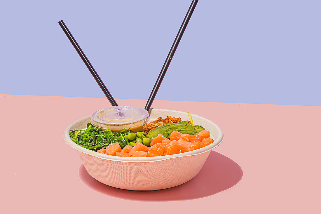Poke-Schale mit frischem Lachs, grünen Edamame-Bohnen, knackigem Algensalat, cremiger Avocado und knusprigem Müsli, dazu eine leichte Dip-Sauce und Stäbchen vor einem zweifarbigen pastellfarbenen Hintergrund