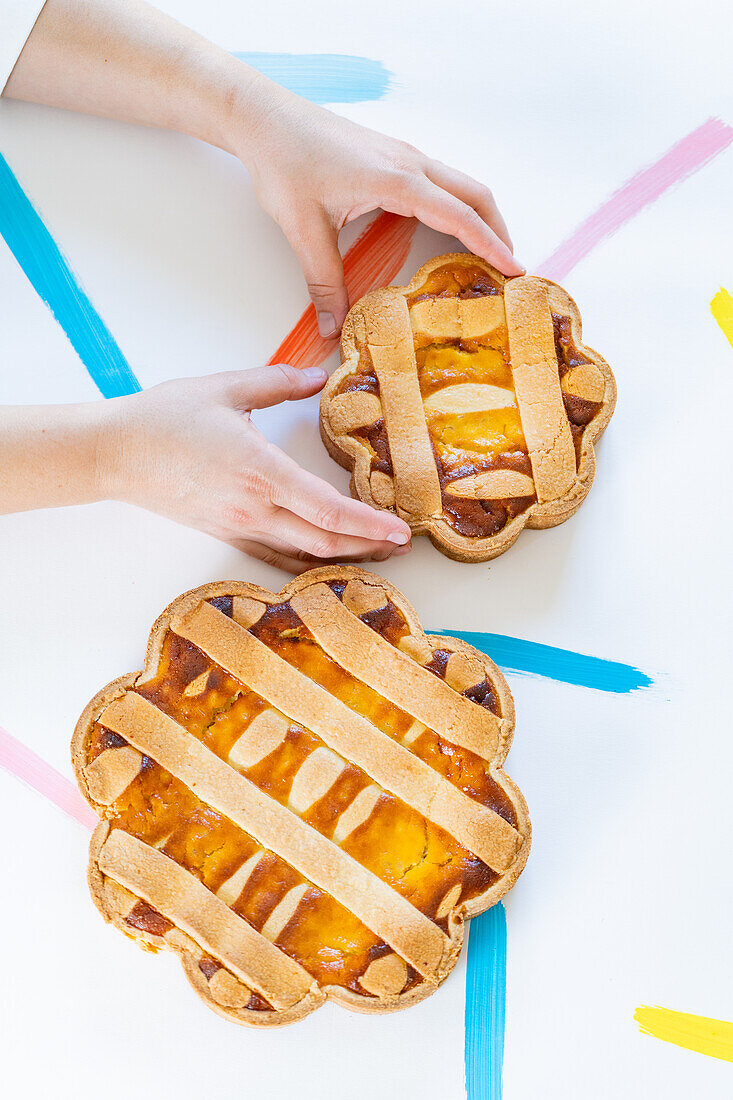 Cropped unrecognizable hands arranging Italian Easter pastiera napoletana pastries on a white surface with colorful brushstrokes.