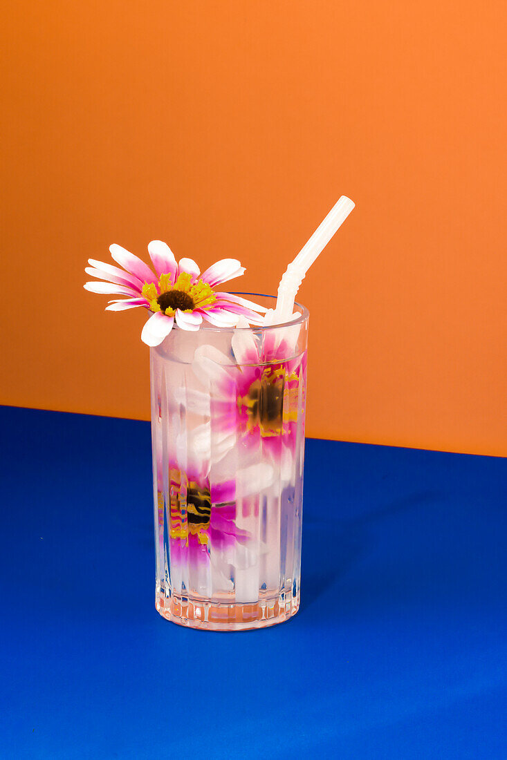 Transparent glass of refreshing cold drink decorated with pink flowers and straw placed on blue surface against bright orange wall