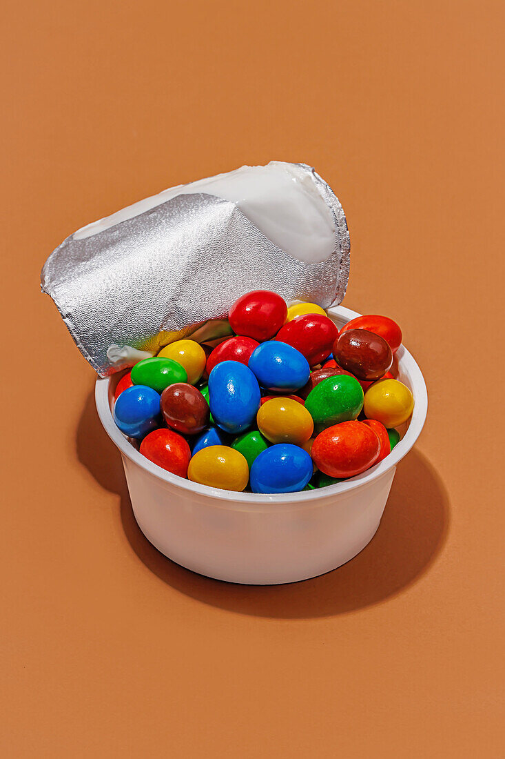 Hoher Winkel des geöffneten Joghurtbehälters, gefüllt mit bunten Bonbons vor orangefarbenem Hintergrund