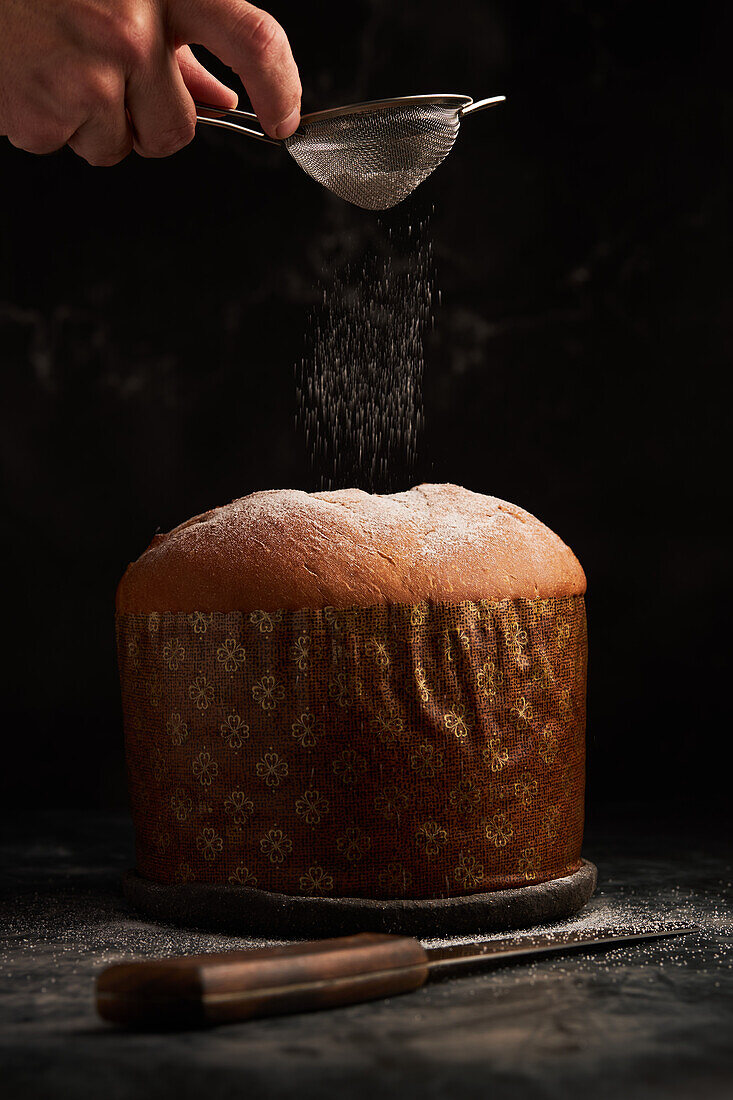 A hand gently dusts powdered sugar onto a panettone, capturing the snow-like sprinkles mid-air against a dark setting