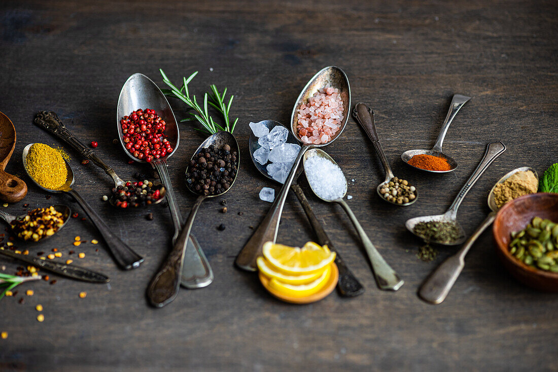 Top view of collection of spoons of various sizes filled with a variety of spices and seeds, such as sunflower seeds, anise star, rosemary, mint, lemon slices, ginger, and assorted peppers, neatly displayed on a dark wooden surface for a rustic appeal.