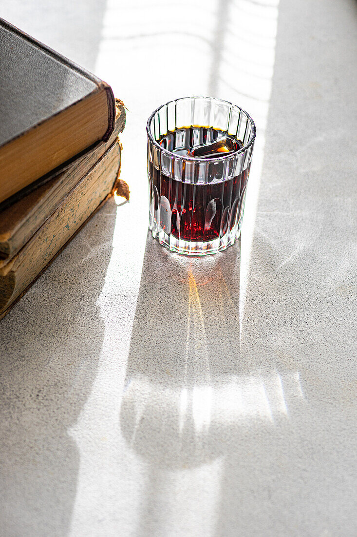 A faceted glass of cherry liqueur casting a long shadow near a pile of old books in sunlight