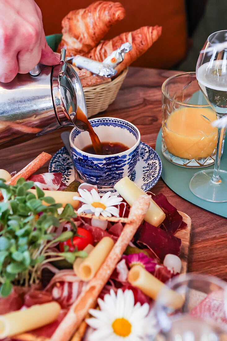 A hand pours coffee into a blue and white cup, part of a gourmet brunch spread including croissants, meats, cheese, and daisies.