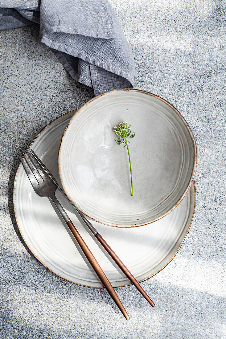 Draufsicht auf einen herbstlich gedeckten Tisch mit Keramikschüssel und -teller mit Blume neben Gabel, Messer und Serviette vor grauem Hintergrund