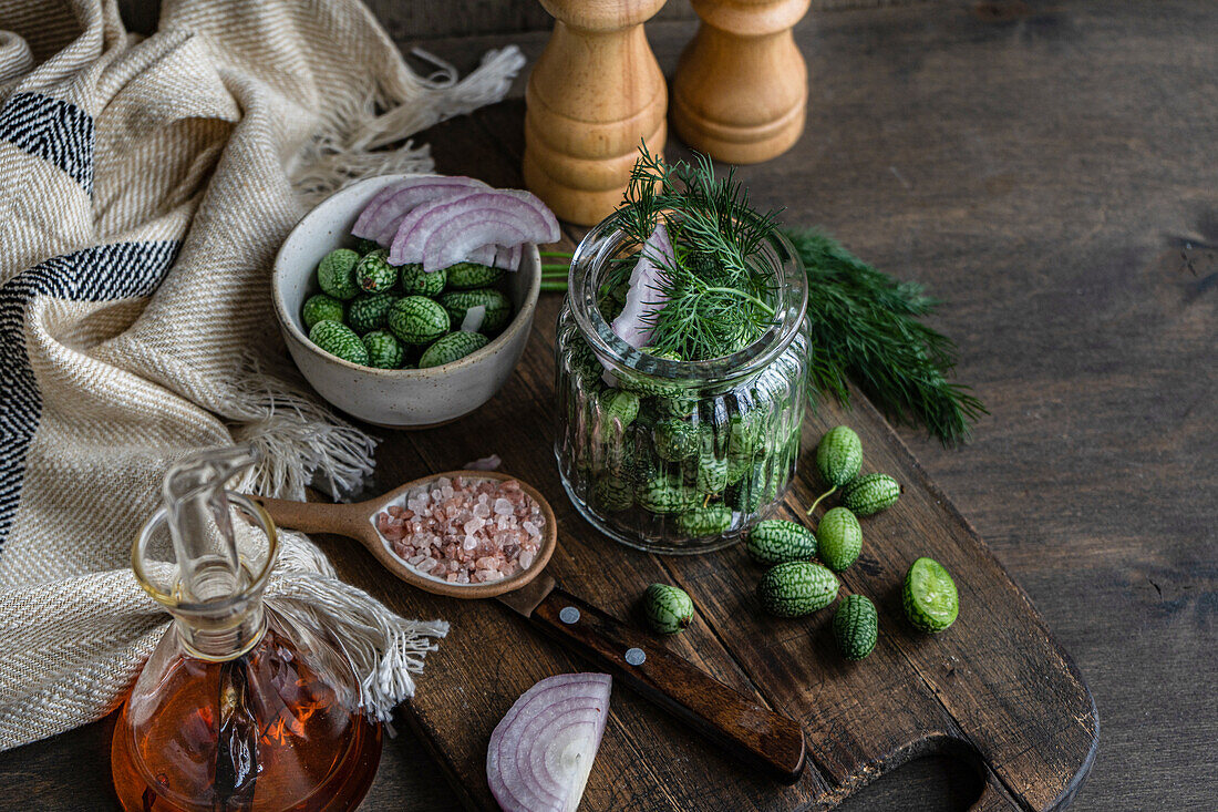 Hochformatige Zutaten für die Zubereitung einer fermentierten Gurke in einem Glas auf einem Holztablett neben einer Serviette und Salz- und Pfefferstreuern vor dunklem Hintergrund