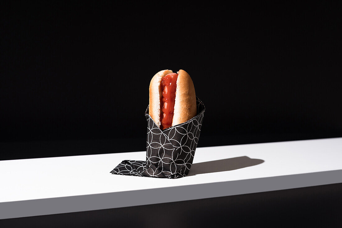 Appetitliches Brötchen mit Wurst und Ketchup auf weißem Holzbrett vor schwarzem Hintergrund im Studio