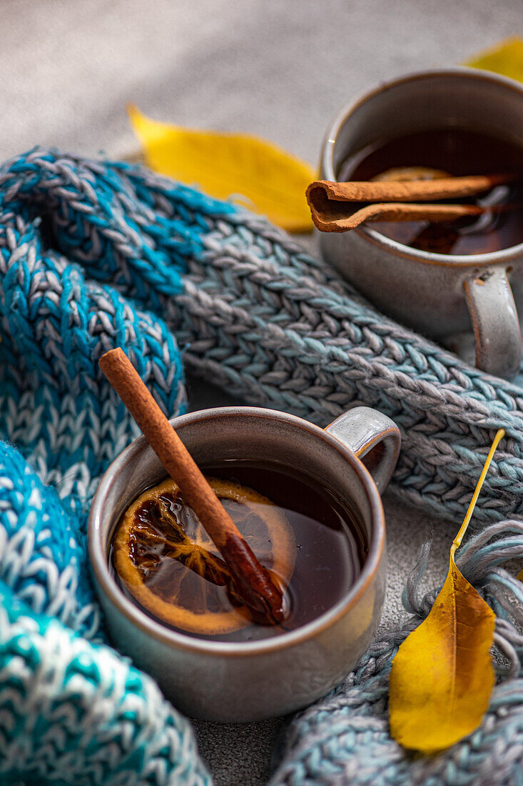 From above of cups aromatic spiced tea with cinnamon sticks and dried orange slices nestled beside a blue knitted fabric complemented by scattered vibrant yellow autumn leaves