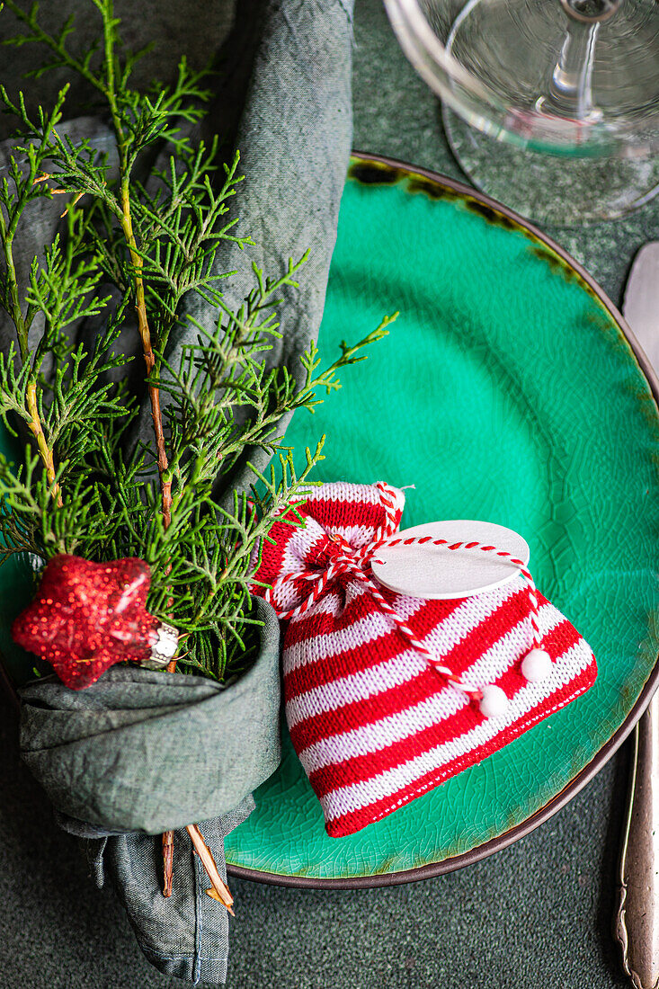 Weihnachtssäckchen und in ein Taschentuch gewickelte Tannenzweige auf einem Teller auf einem grünen Tisch neben Glas und Messer
