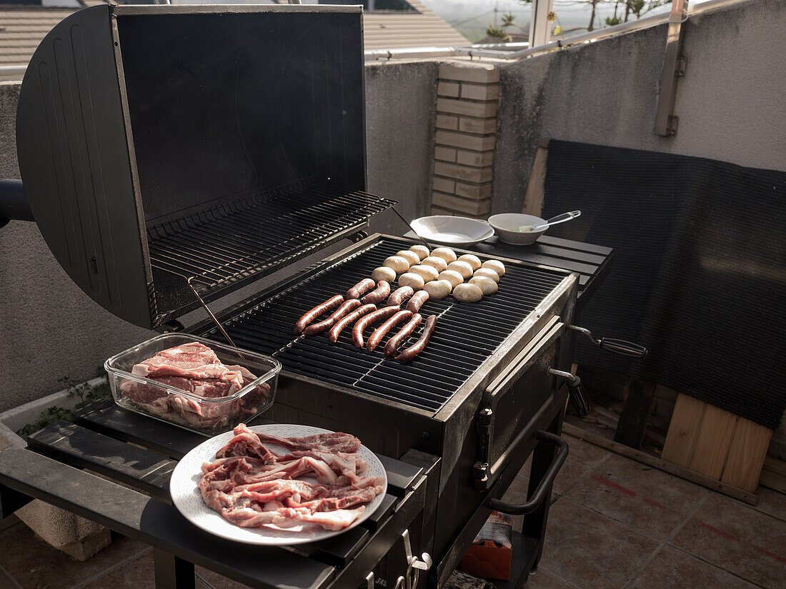 High angle of fresh raw meat placed in plate and glass container on wooden planks while mushrooms sausages arranged neatly on barbecue grill rack in rooftop kitchen