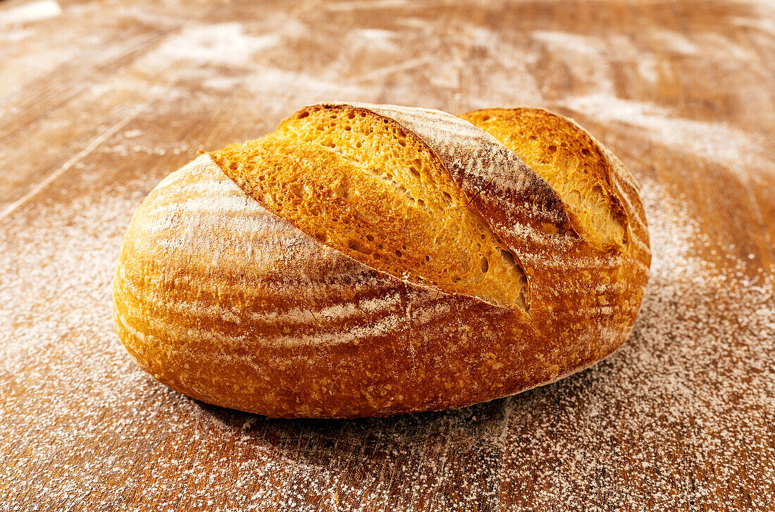 Nahaufnahme eines frisch gebackenen, leckeren Brotes mit knuspriger Kruste auf einem Holztisch mit Mehl rundherum in der Küche einer Bäckerei