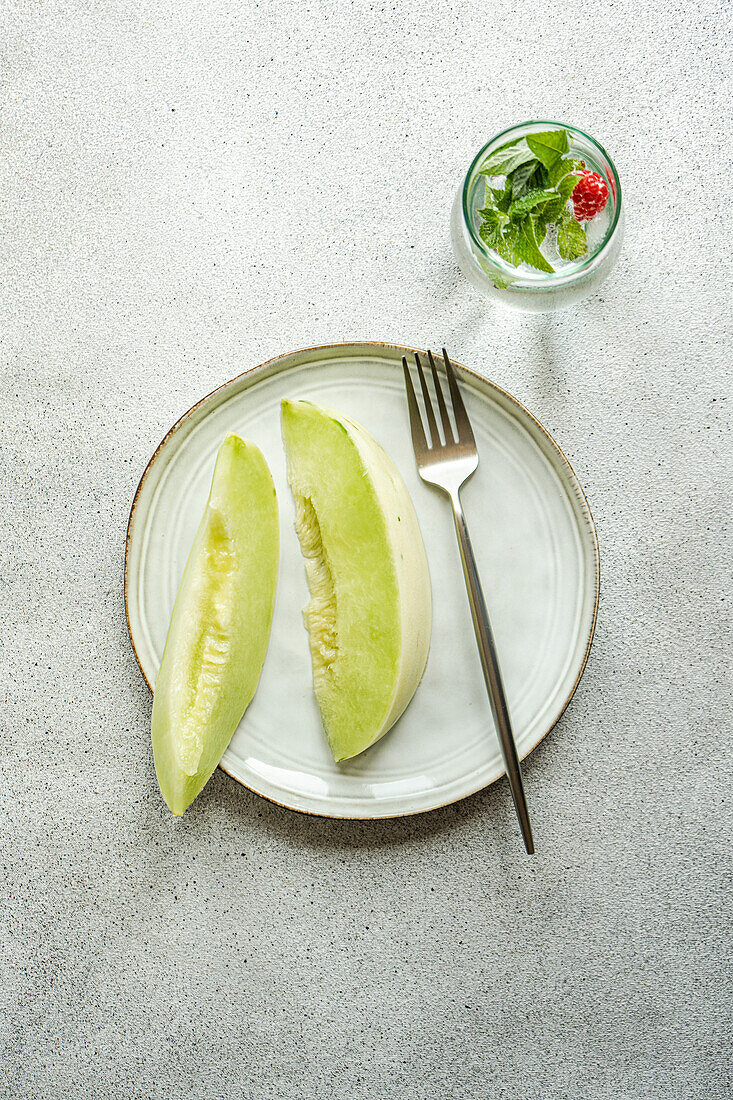 From above slices of green melon placed in a ceramic dish with a lemonade on concrete background