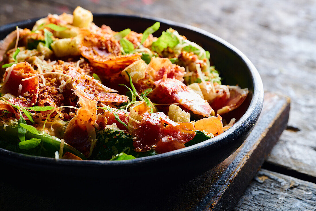 Hoher Winkel von appetitlichen Salat, bestehend aus frischem gehackten Gemüse, Kräutern und Speck in einer schwarzen Schüssel serviert
