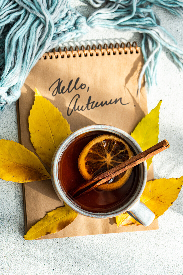 Top view of cup spiced tea with cinnamon sticks and dried orange slices, surrounded by vibrant yellow autumn leaves are juxtaposed against a notebook inscribed with "Hello Autumn!" on a textured gray background
