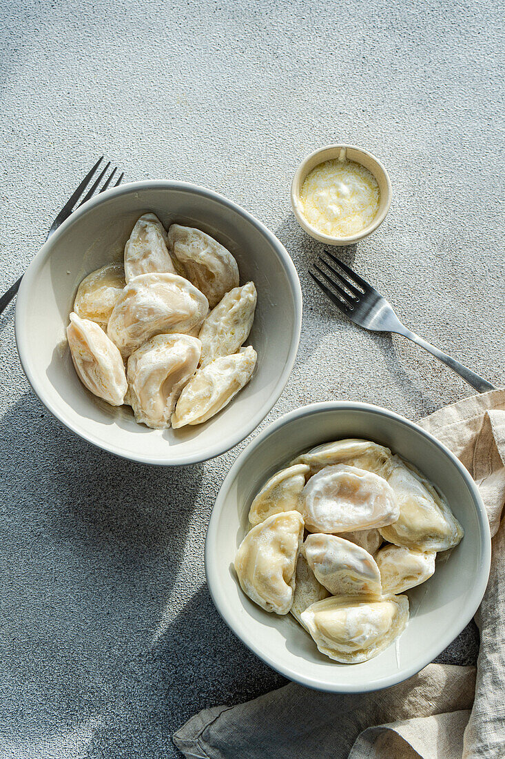 From above of overhead view of Ukrainian varenyky dumplings in two white bowls with a side of sour cream, on a light textured surface, accompanied by a linen napkin and a fork