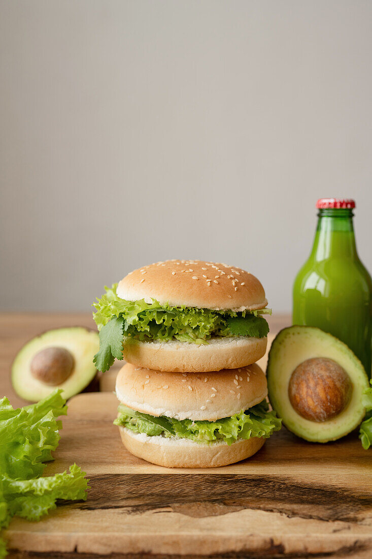 Tasty vegetarian burgers with fresh lettuce near avocado and bottle of juice on wooden cutting board against gray background