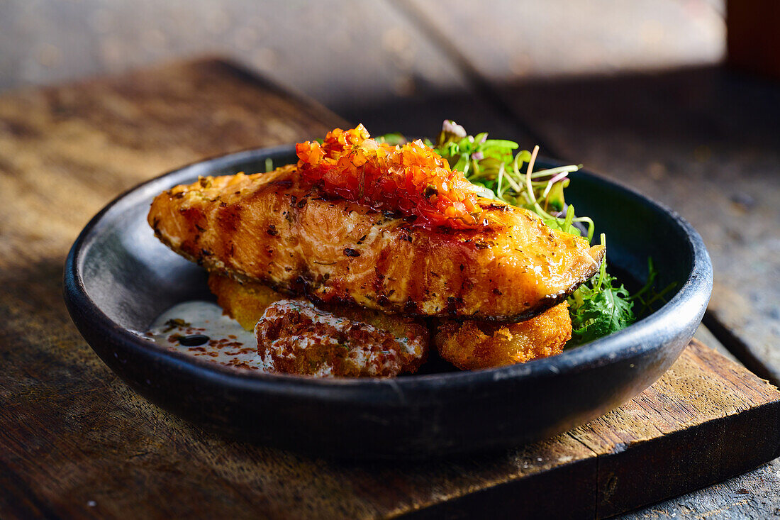 High angle of appetizing grilled salmon steak garnished with sauce and herbs served on black bowl placed on wooden cutting board