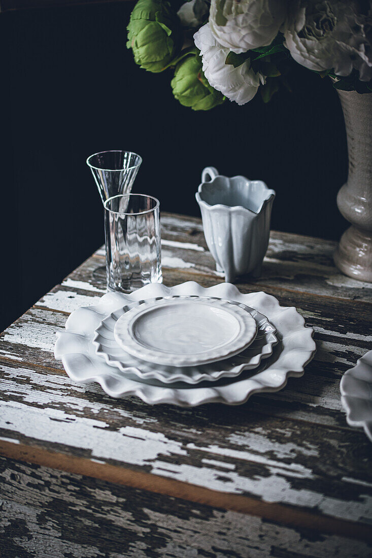 Weiße Keramikteller stehen neben Glaswaren und einem Strauß frischer Blumen auf einem schäbigen Holztisch