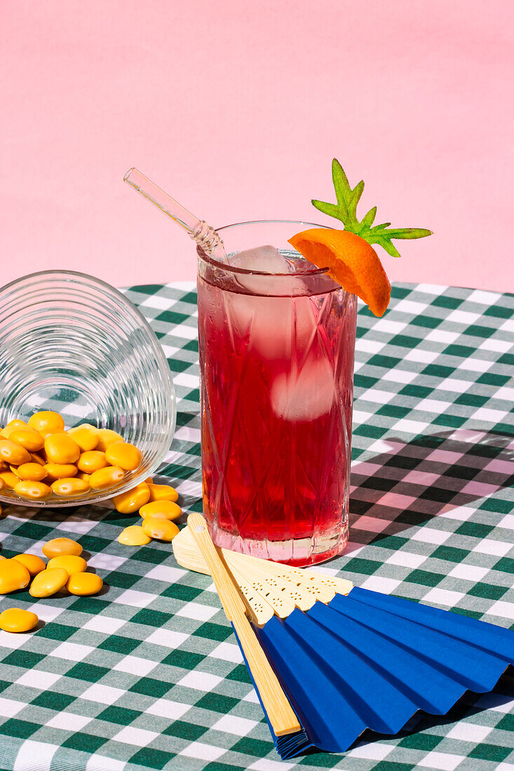 Glass of cold refreshing red cocktail with ice and straw served with orange near transparent bowl with lupin beans and fan on table with checkered tablecloth