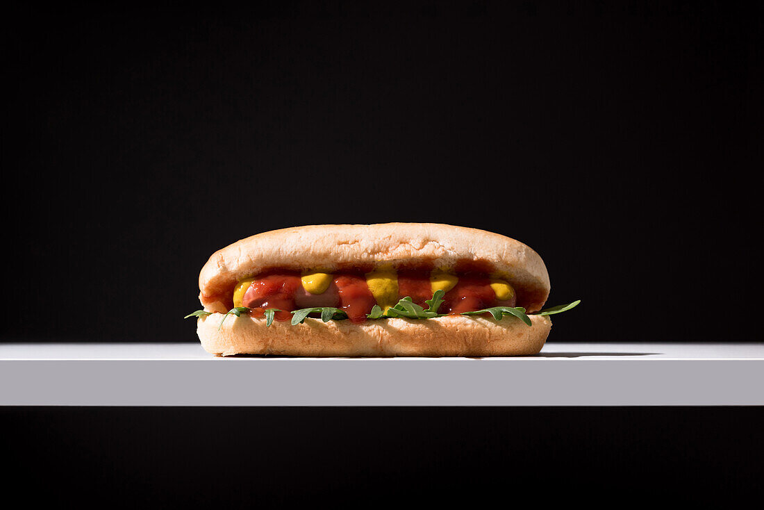 Appetizing bun with sausage and ketchup with rocket salad served table mat over white wooden board against black background in studio