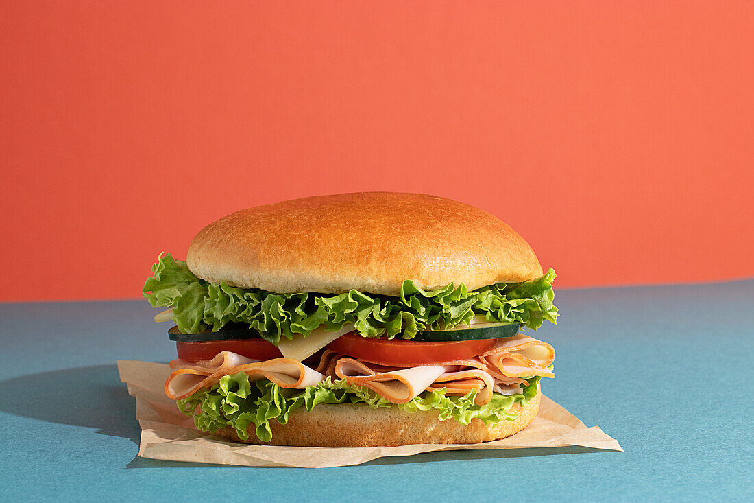 Leckeres Sandwich mit Schinken, Tomaten, Gurken- und Käsescheiben und frischem Salat auf blauem und orangem Hintergrund