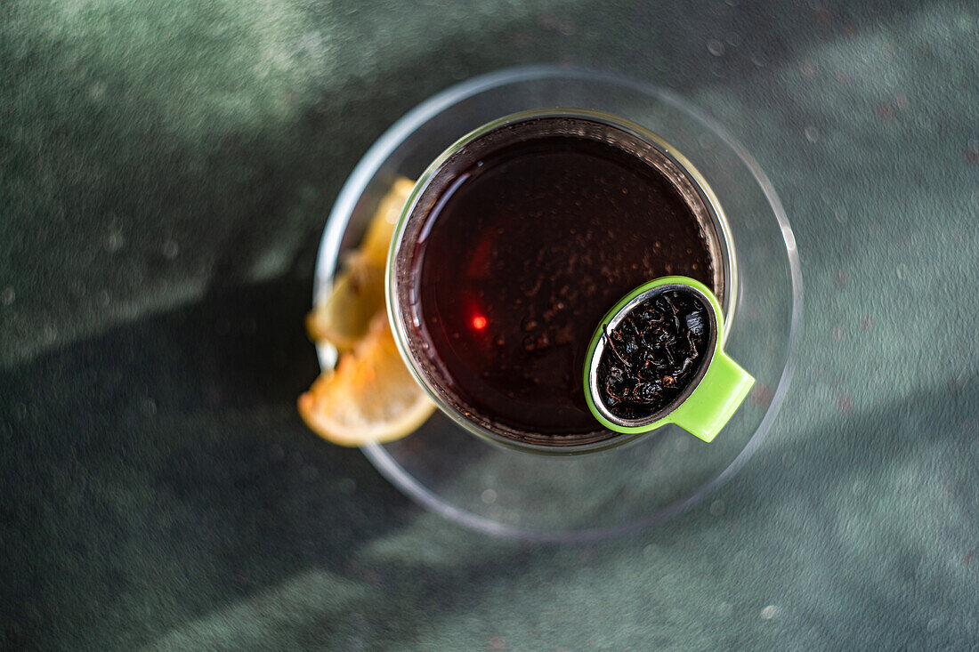 Close up of clear glass of tea with a slice of lemon and a green tea strainer on a textured dark background